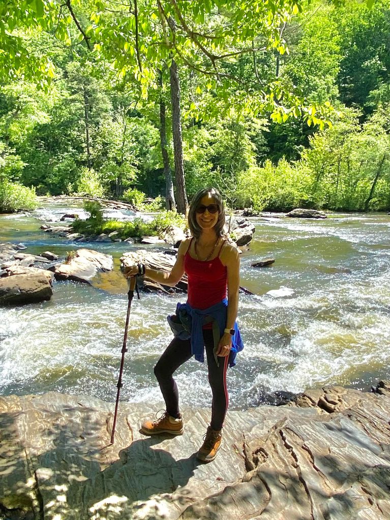 Adaris Mas holding a hiking stick standing on rocks next to a rushing river.